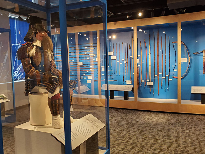 A Samurai suit of armor sits on display in the middle of the exhibit of other weapons. 