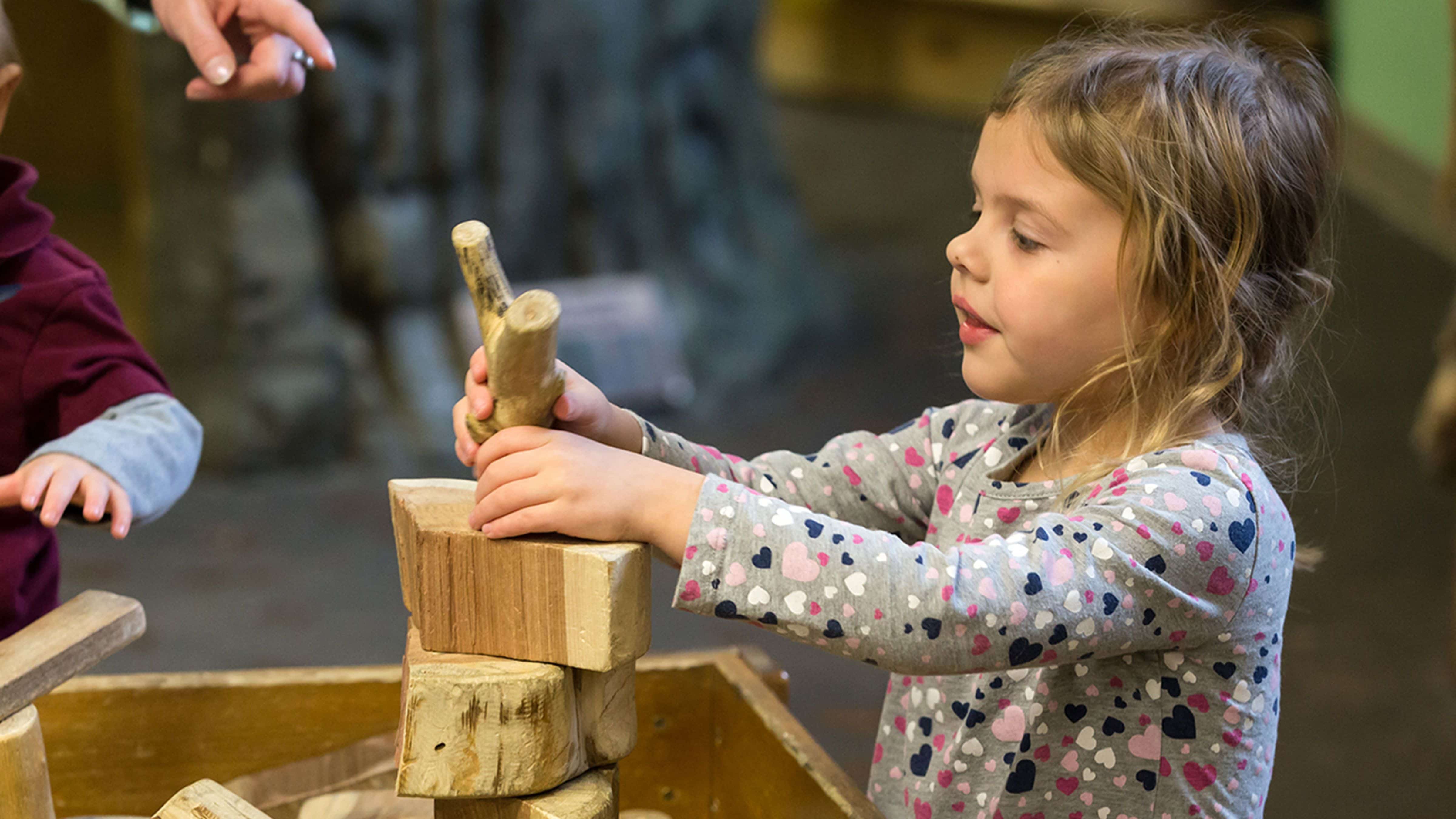 Little girl playing building structure with play wooden stick and blocks, adult pointing at stick out-of-frame