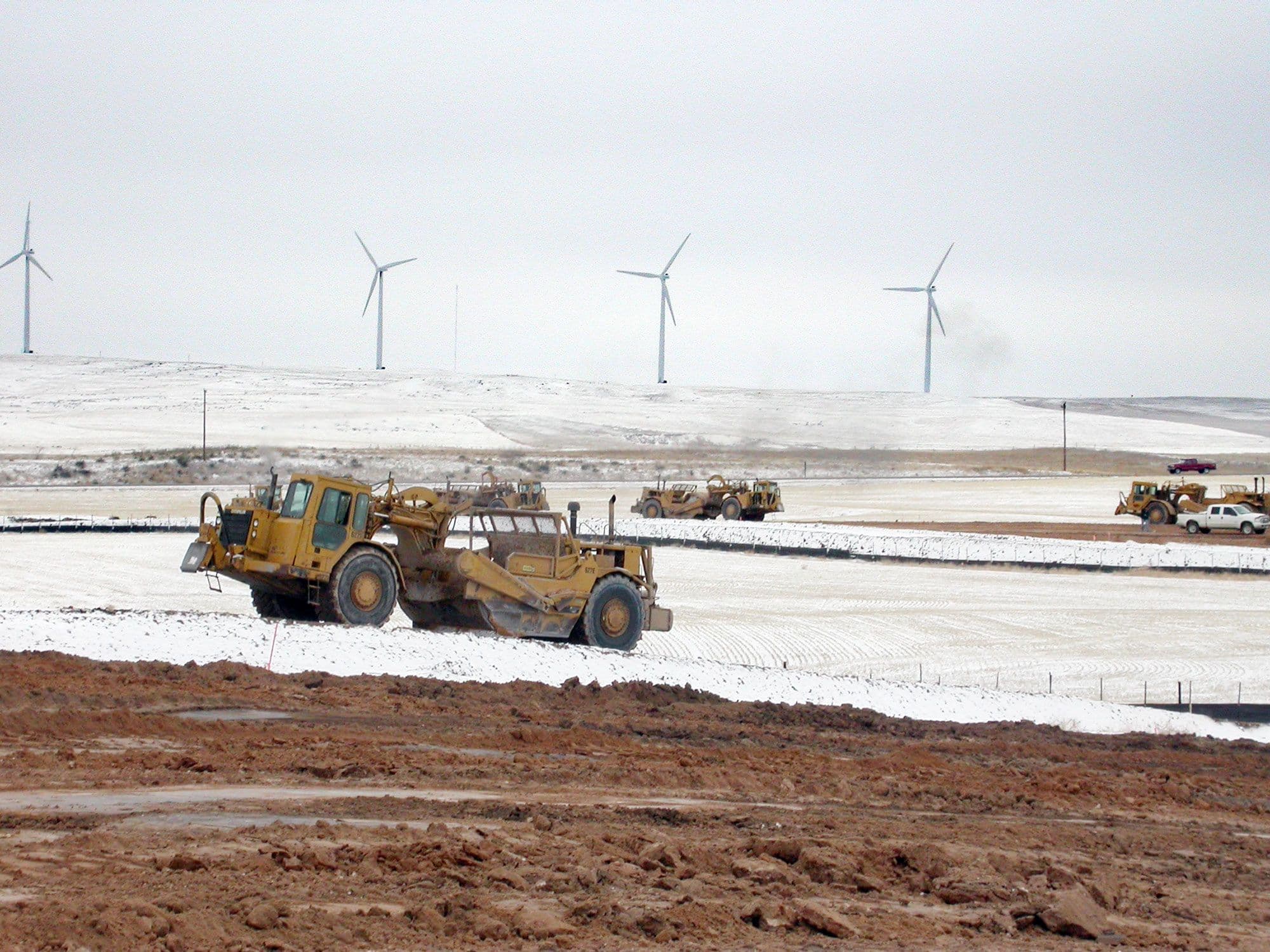 Heavy construction machinery travels across snowy land near road construction with wind turbines in the background.
