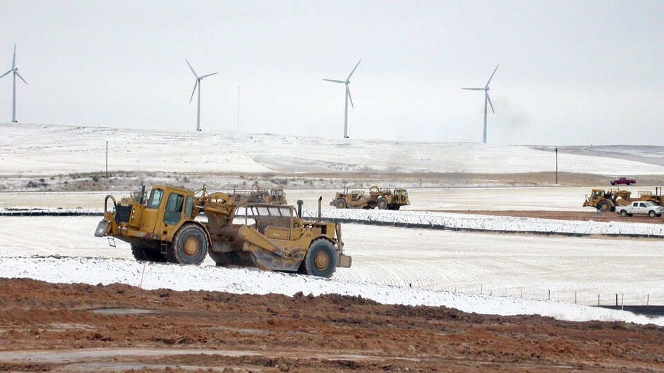 Heavy construction machinery travels across snowy land near road construction with wind turbines in the background.