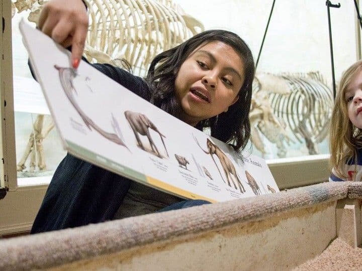 A woman holds up a book with illustrations of animals up as she explains them to a small group of children.